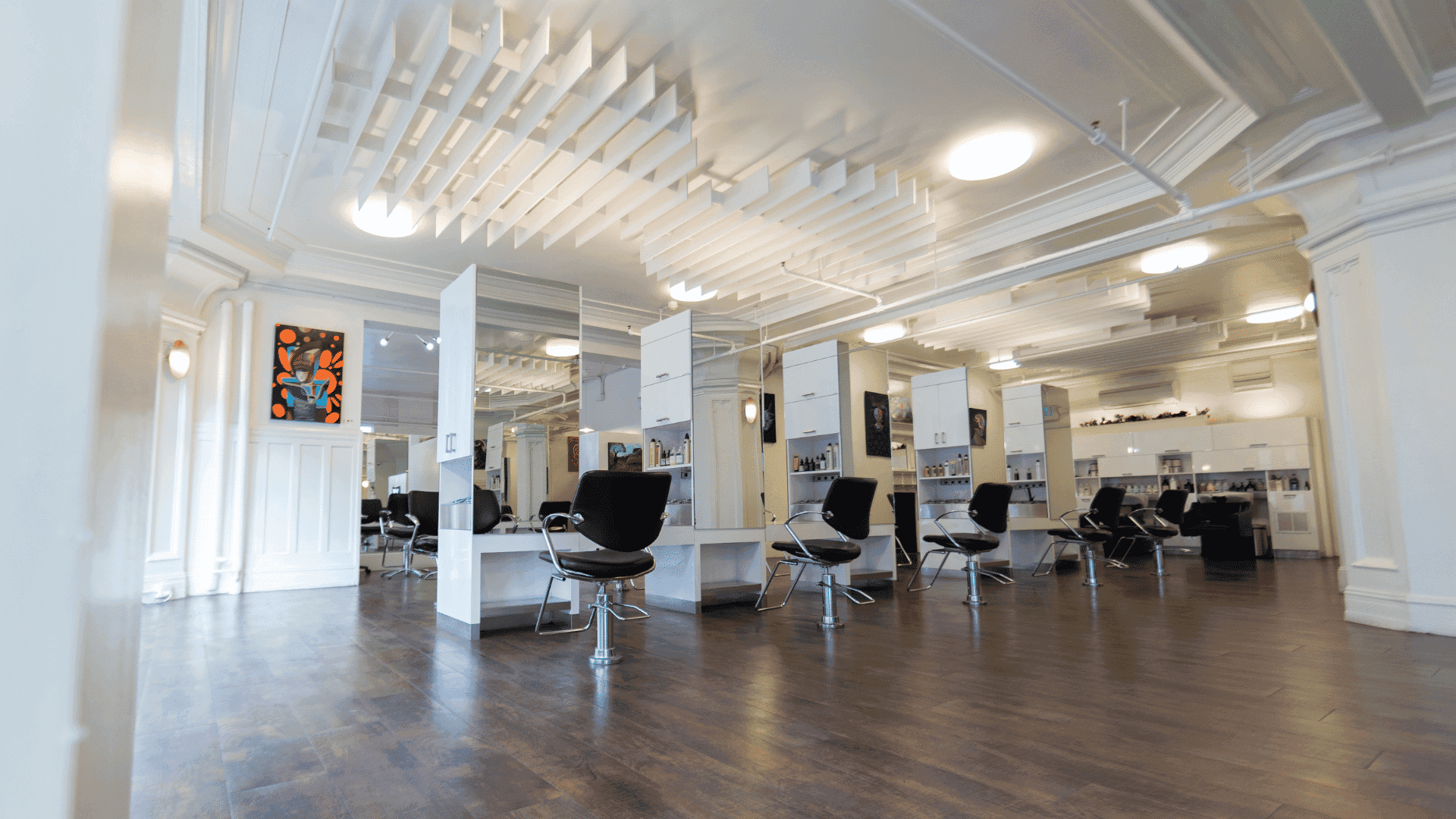 Modern, spacious salon with black chairs, white stations, and wooden floors. Bright and stylish setup.